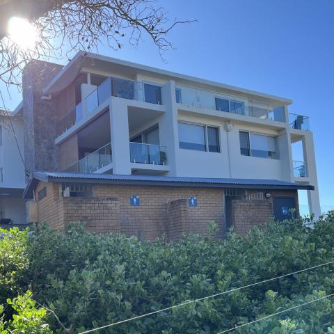 Existing facility building at Karagi Point Reserve, The Entrance North Old brown bricked toilet building with gabled roof. Apartment building in the background. Shrub in the foreground.