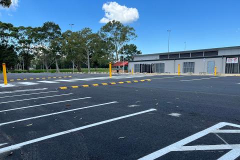 Rogers Park Woy Woy car park Car park with white markings, yellow and black speed bumps, yellow parking poles, sports amenities building and oval and trees in the background.
