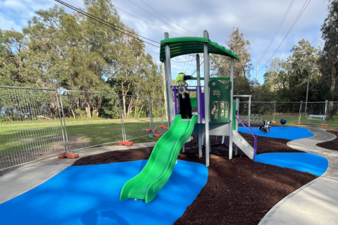 Toukley Woodland playspace Photo of playspace with blue softfall, a green and grey play unit with a green slide, a concrete pathway and bush in the background